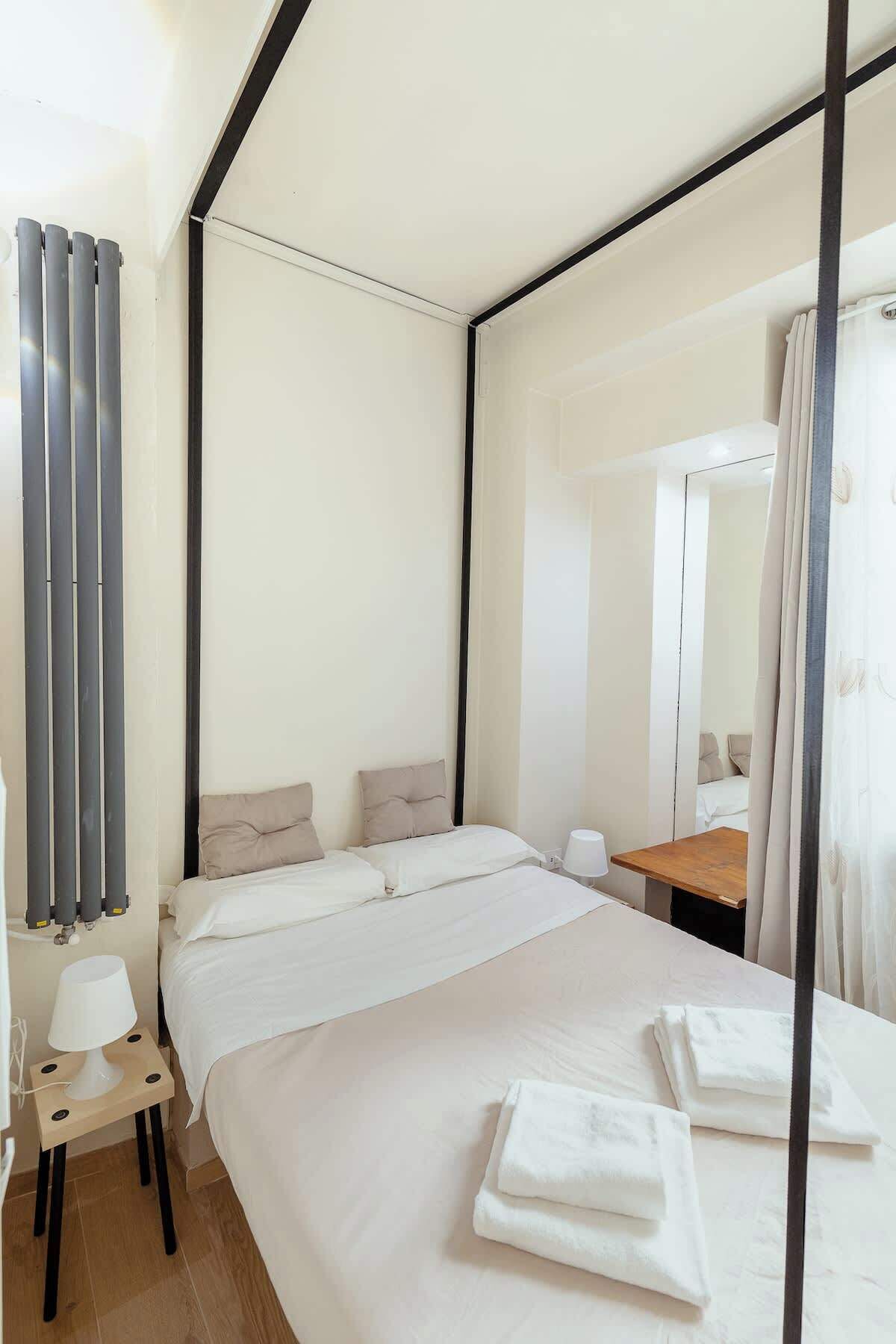 Ein modernes Hotelzimmer mit einem großen Bett, einem Nachttisch, einer Lampe, einem Spiegel und einem Fenster mit Vorhängen.