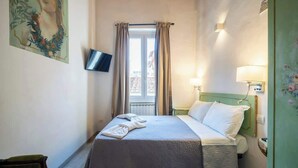 A hotel room with a bed, a television, a window with curtains, a lamp, and a painting on the wall. - Duomo Studio Room with Private Bath and Kitchenette (Firenze)