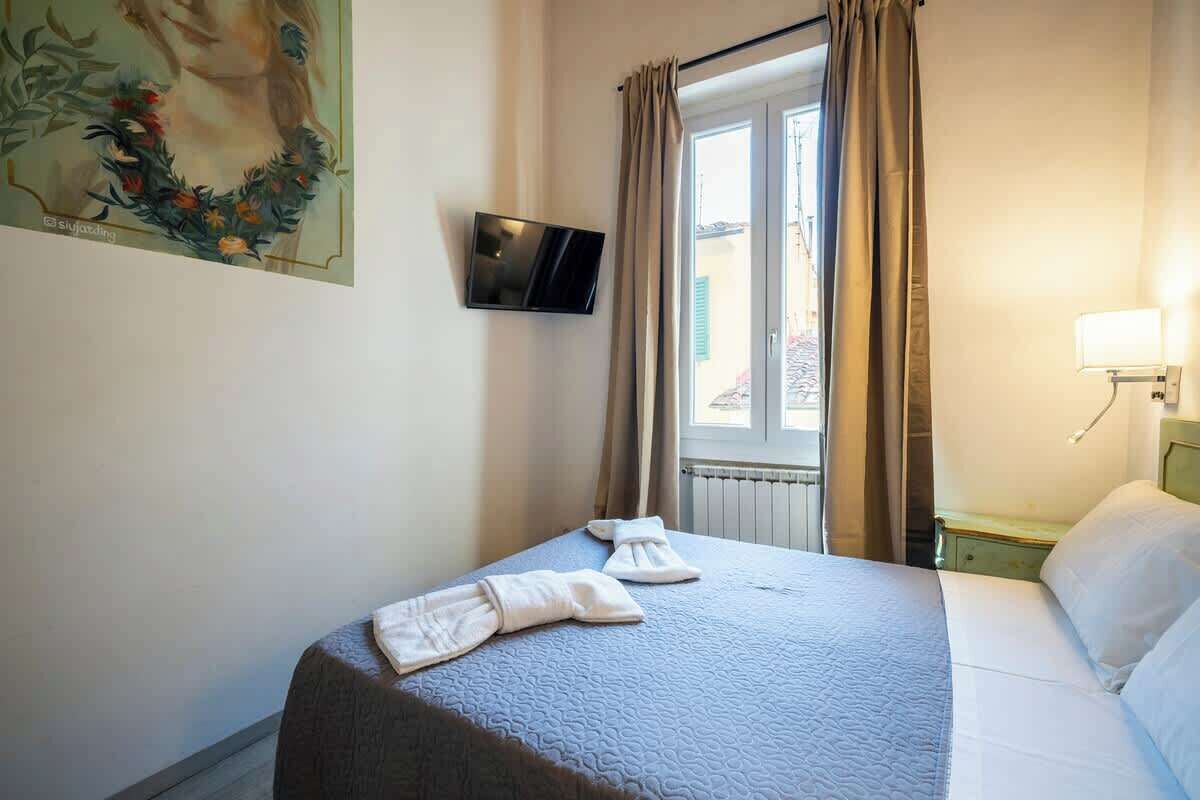 A hotel room with a bed, a flat-screen TV mounted on the wall, a window with curtains, and a painting on the wall.