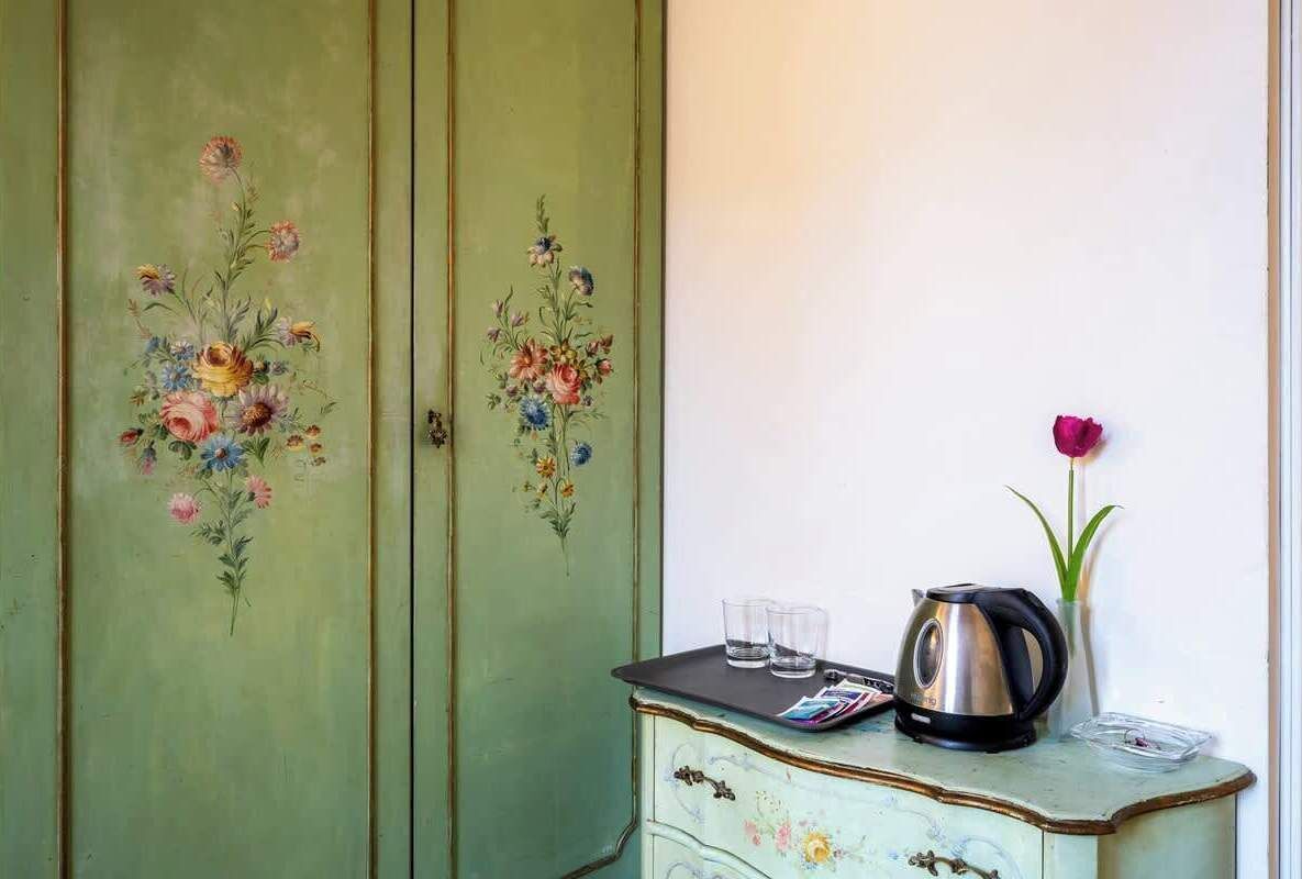 Een antieke groene dresser met bloemmotieven, een waterkoker en een vaas met daarin een enkele roos.