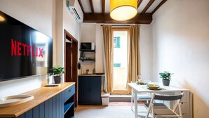 Quarto compacto com bancada de madeira, TV exibindo "NETFLIX", pequena cozinha, área de refeições com mesa e cadeiras, e janela com cortinas.