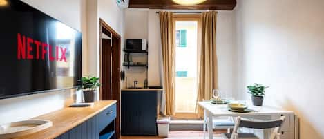 A compact room with a wooden counter, a TV displaying "NETFLIX," a small kitchenette, a dining area with a table and chairs, and a window with curtains.
