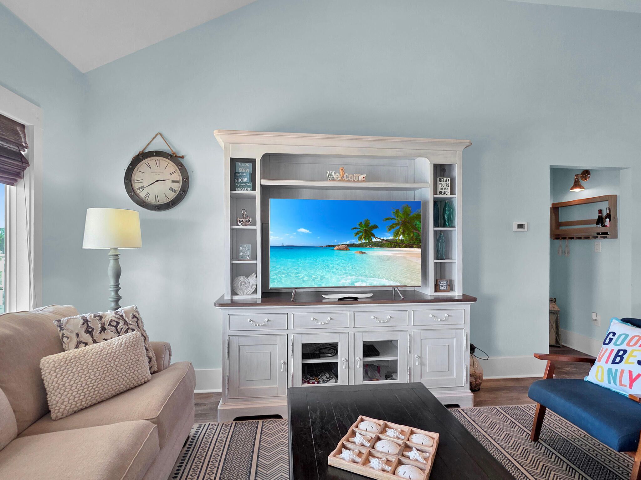 A living room with a television displaying a beach scene, a clock on the wall, and a sofa with decorative pillows.