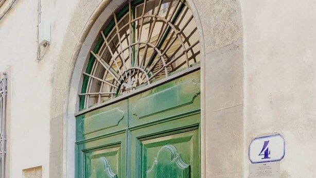 Une porte à deux battants, peinte en vert, ornée de poignées décoratives, une grille métallique au-dessus, et une plaque fixée au mur, sur le côté droit.