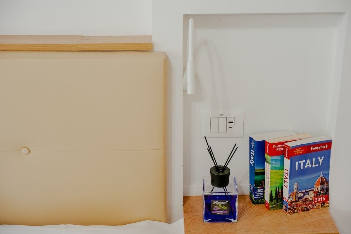 Ein Bett mit Holz-Kopfstück, ein kleiner Tisch mit einer Glasflasche und Büchern, sowie eine Wandsteckdose.