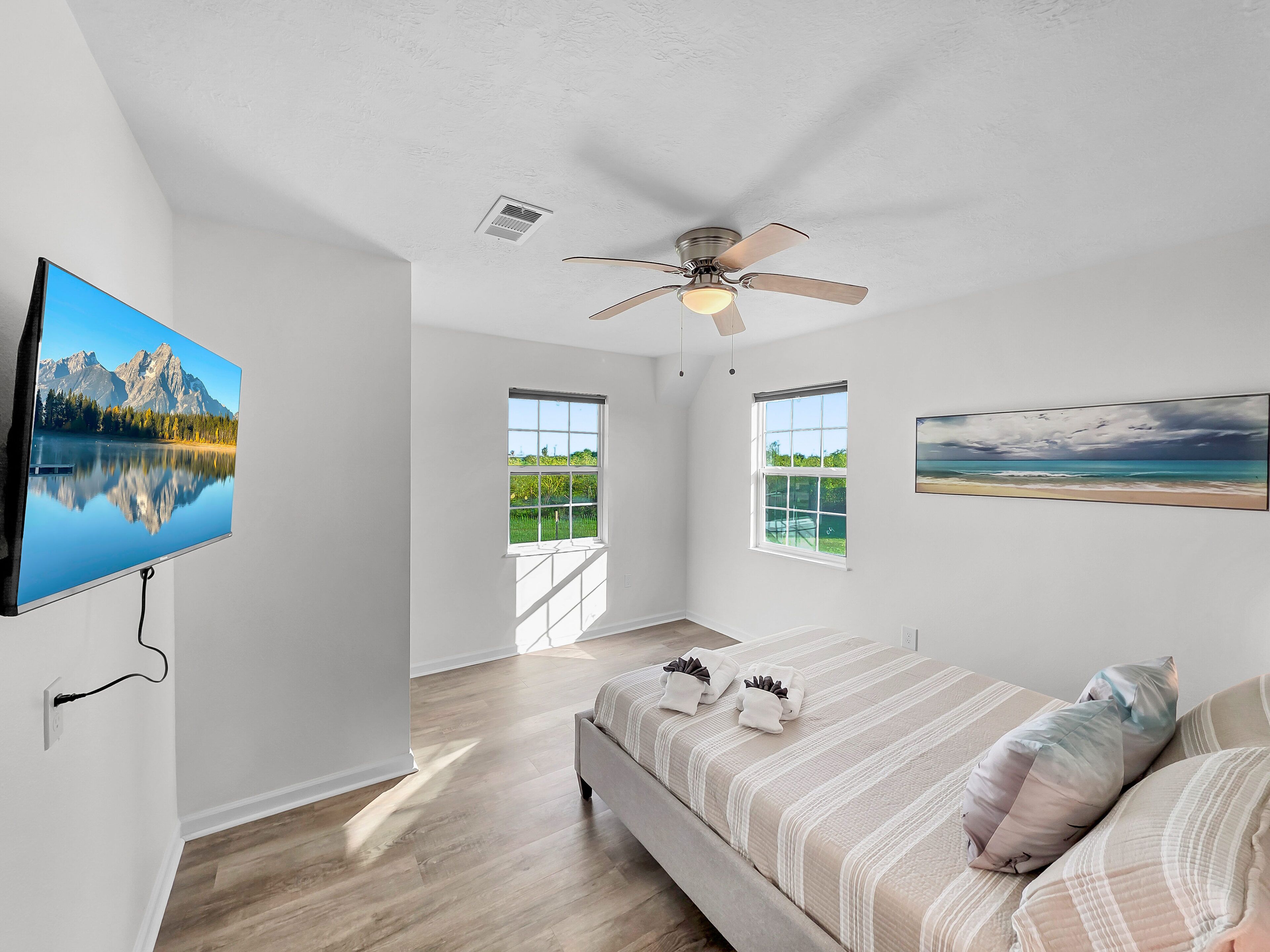 A spacious bedroom with a large bed, two windows, a ceiling fan, and a TV on the wall.