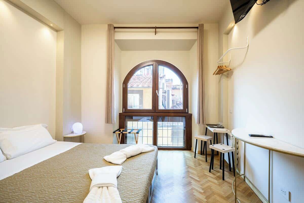 Une chambre d’hôtel avec un grand lit, une petite table, deux chaises, un bureau et une grande fenêtre donnant sur des immeubles.