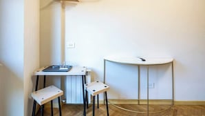 A small, modern room with a white radiator, a white table with a laptop, two stools, a white sink, and a wooden hanger on a white rod. - Duomo Room with Private Bath, WiFi and Netflix (Firenze)