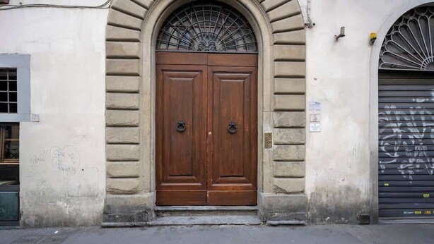 Une porte en bois à deux battants, surmontée d’une grille métallique, est encastrée dans un mur de pierre, avec une petite fenêtre sur le côté gauche.