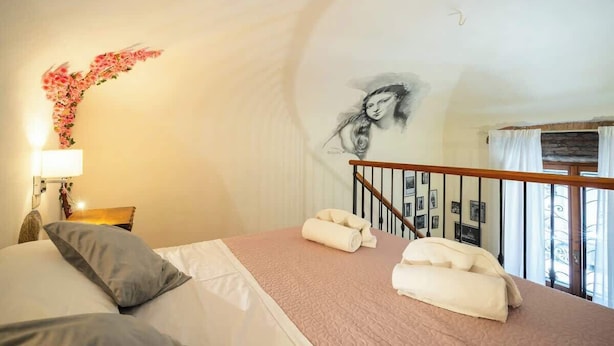 Een slaapkamer met een bed, kussens, een nachtkastje, een lamp, een aan de muur hangend kunstwerk en een raam met gordijnen.
