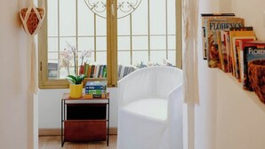A room with a large arched window, a white chair, a small table with books, and a wall-mounted shelf with books.