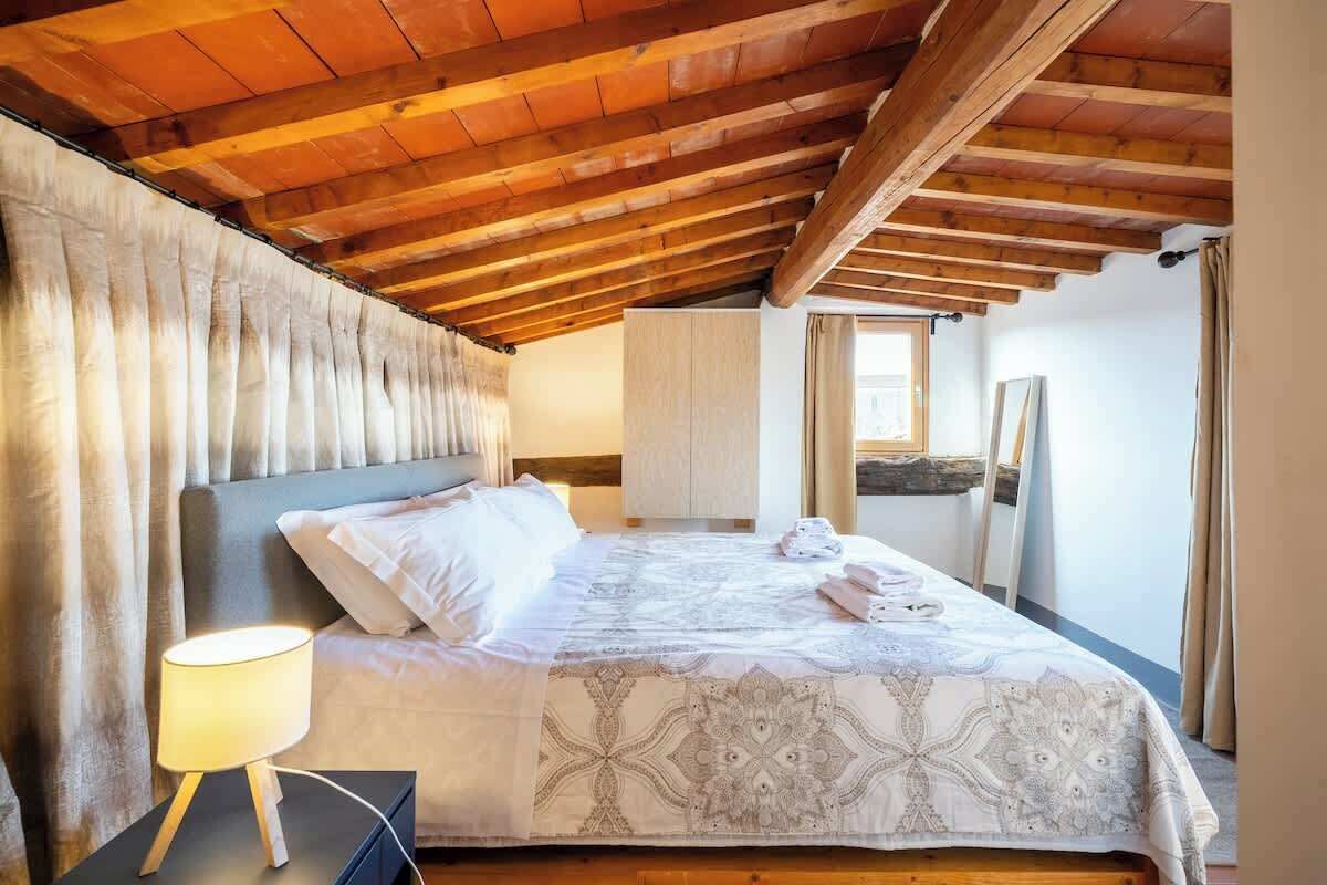 Un dormitorio con techo de madera, una cama con colcha estampada, una lámpara de noche y una ventana con cortinas.