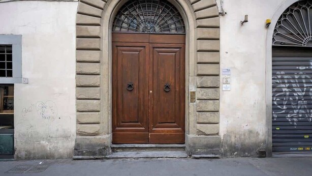 Une porte en bois à deux battants, surmontée d’une grille métallique décorative, est encastrée dans un mur de pierre, avec une petite fenêtre sur le côté gauche.