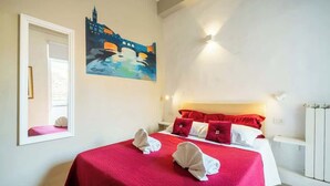 A hotel room with a bed, red and white bedding, a painting on the wall, and a window with a view.