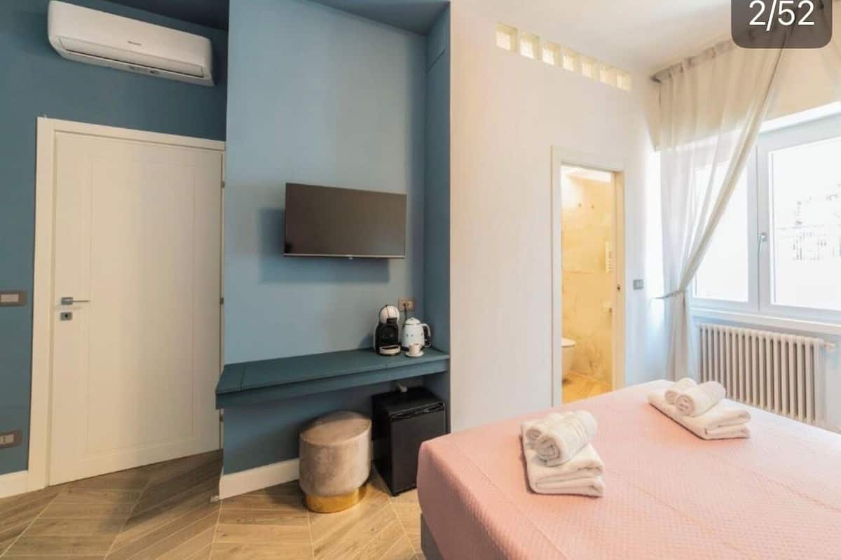 A modern hotel room with a flat-screen TV, a small seating area with a coffee machine, a bed with towels, and a window with curtains.