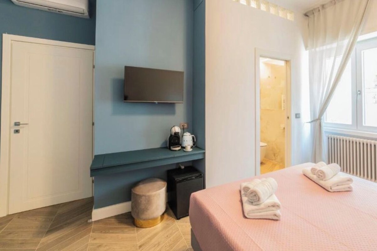 A modern hotel room with a flat-screen TV, a small seating area with a coffee machine, a bed with towels, and a window with curtains.