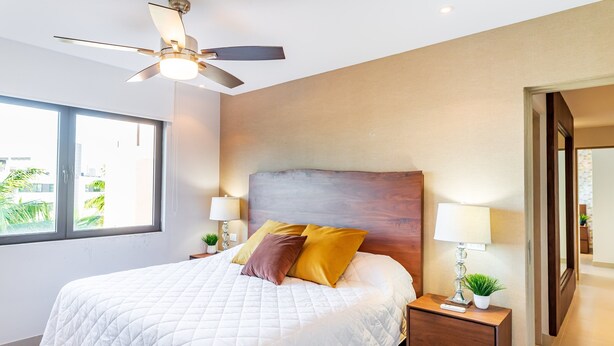 A bedroom with a large bed, two bedside lamps, a nightstand, a ceiling fan, and a view of greenery outside. - D410 Lorena Ochoa 4 Bedroom Apts (Playa del Carmen)