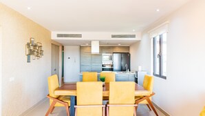 A modern dining area with a wooden table and yellow chairs, a kitchen in the background, and a beige textured wall. - D410 Lorena Ochoa 4 Bedroom Apts (Playa del Carmen)