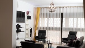 Un salon moderne avec une table basse en verre, deux fauteuils en cuir noir, un canapé noir, un téléviseur fixé au mur et un lustre.