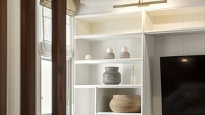 A modern living room with a flat-screen TV mounted on the wall, a grey ottoman, and a decorative shelf with various items. - Charming Family Retreat – 4BR Marylebone (Greater London)