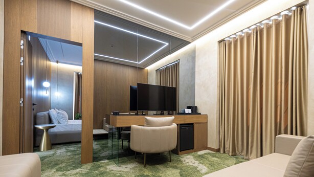A modern hotel room with a sofa, a desk with a TV, a small table, and a green patterned carpet. - Sazani Hotel (Vlorë)