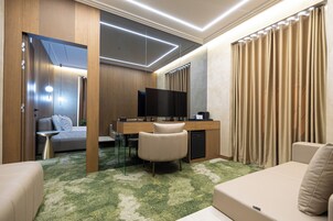 Una habitación de hotel moderna con un sofá, un escritorio con televisor, una mesita pequeña y una alfombra verde estampada.