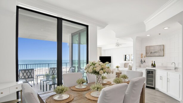 A modern kitchen with white cabinetry, a dining area with a wooden table and white chairs, and a view of the ocean through large glass doors. - Phoenix Ix 307 2 Bedroom Condo (Orange Beach)