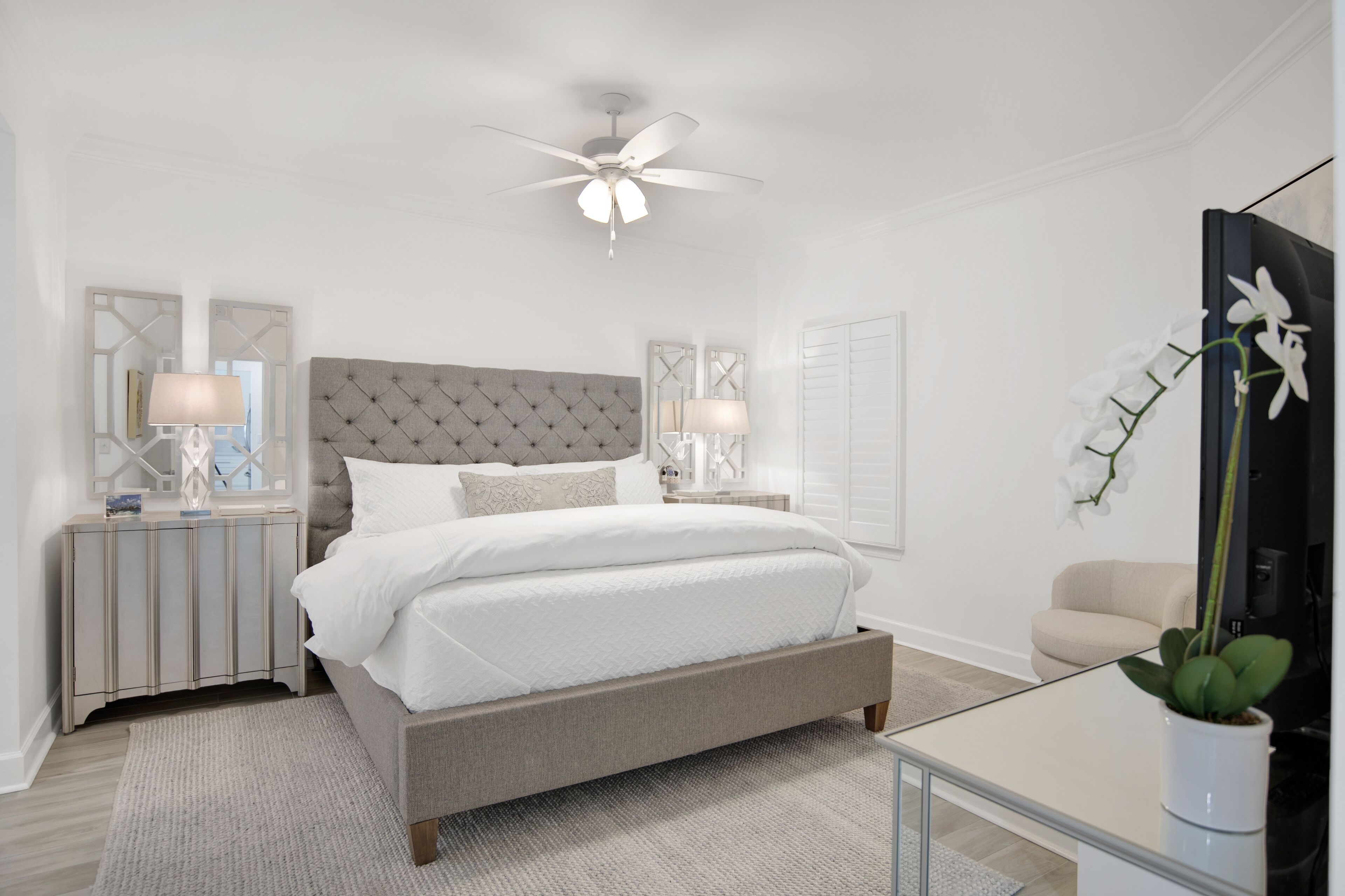 A bedroom with a tufted headboard, a bed with white bedding, two bedside tables with lamps, a ceiling fan, a potted orchid, and a television on a stand.