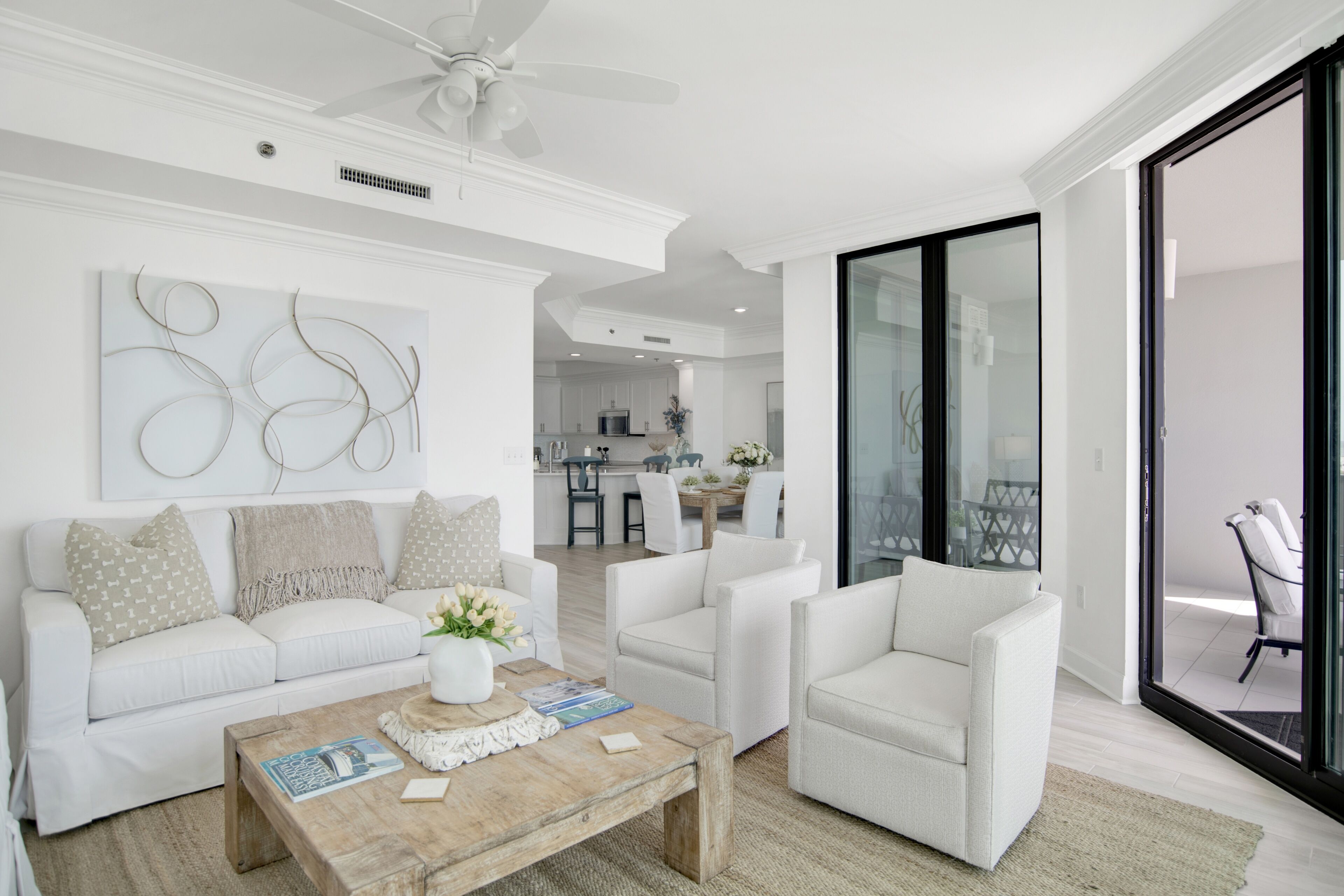 Un salon moderne avec un canapé blanc, des fauteuils et une table basse en bois. On aperçoit un ventilateur de plafond et une cuisine à l’arrière-plan.