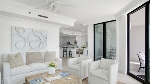 A modern living room with a white sofa, armchairs, and a wooden coffee table. A ceiling fan and a kitchen are visible in the background. - Phoenix Ix 307 2 Bedroom Condo (Orange Beach)