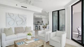 Un salon moderne avec un canapé blanc, des fauteuils et une table basse en bois. On aperçoit un ventilateur de plafond et une cuisine à l’arrière-plan.