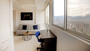 A modern living room with a sofa, a coffee table, and a desk with a view of a cityscape through large windows. - Apartamentos Oslo-by Medellin Experience (Medellin)