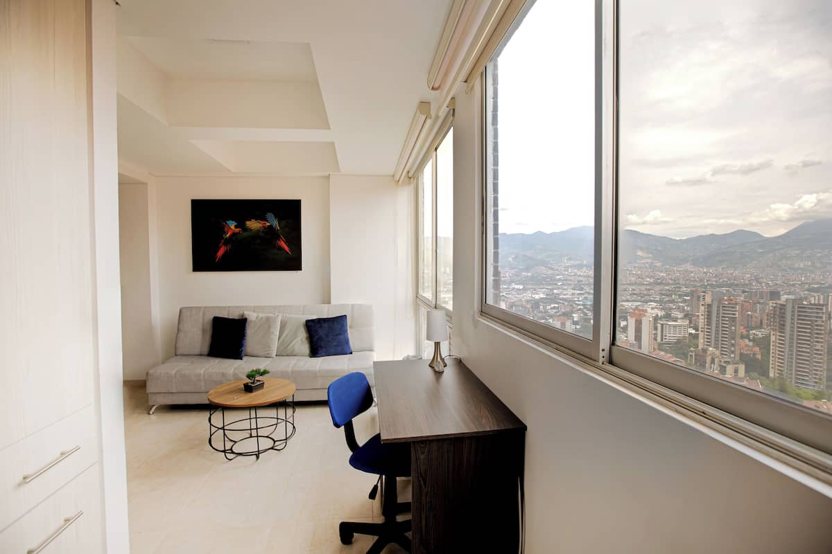 A modern living room with a sofa, a coffee table, and a desk with a view of a cityscape through large windows.