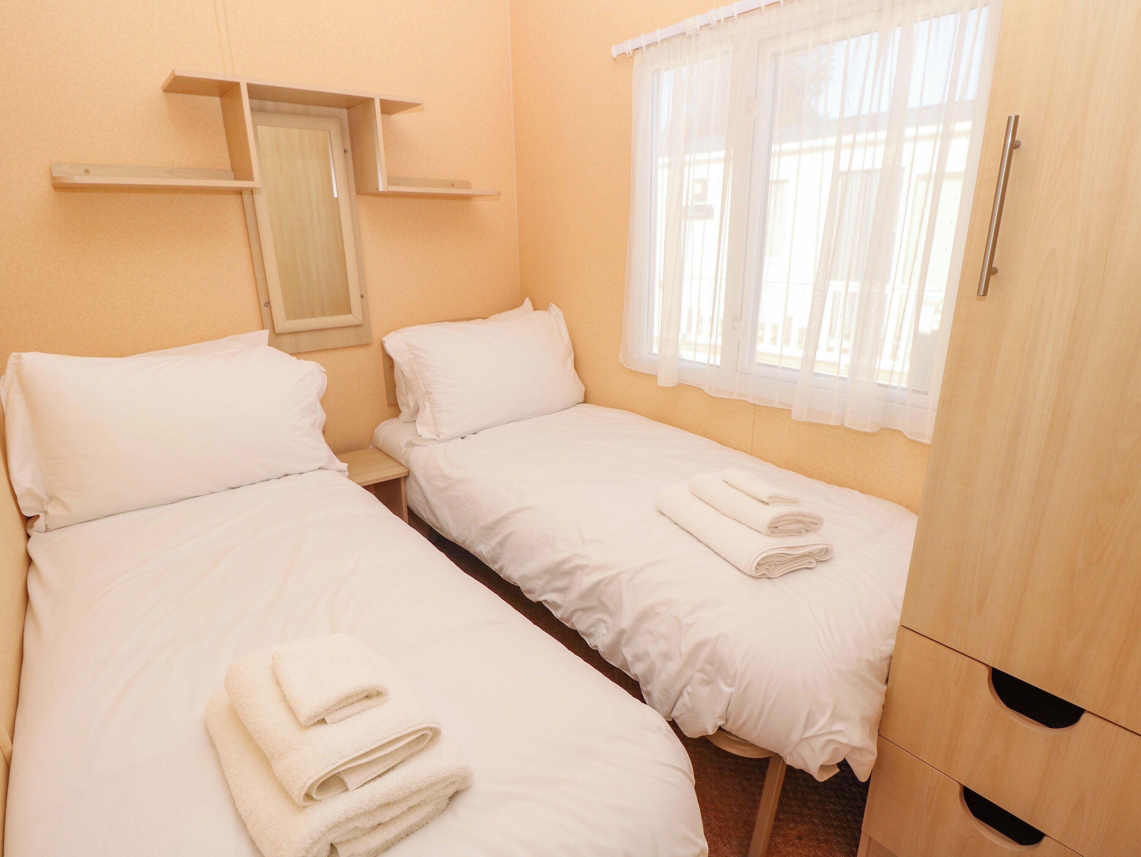 A hotel room with two single beds, a wooden wardrobe, a window with sheer curtains, and a small shelf above the door.