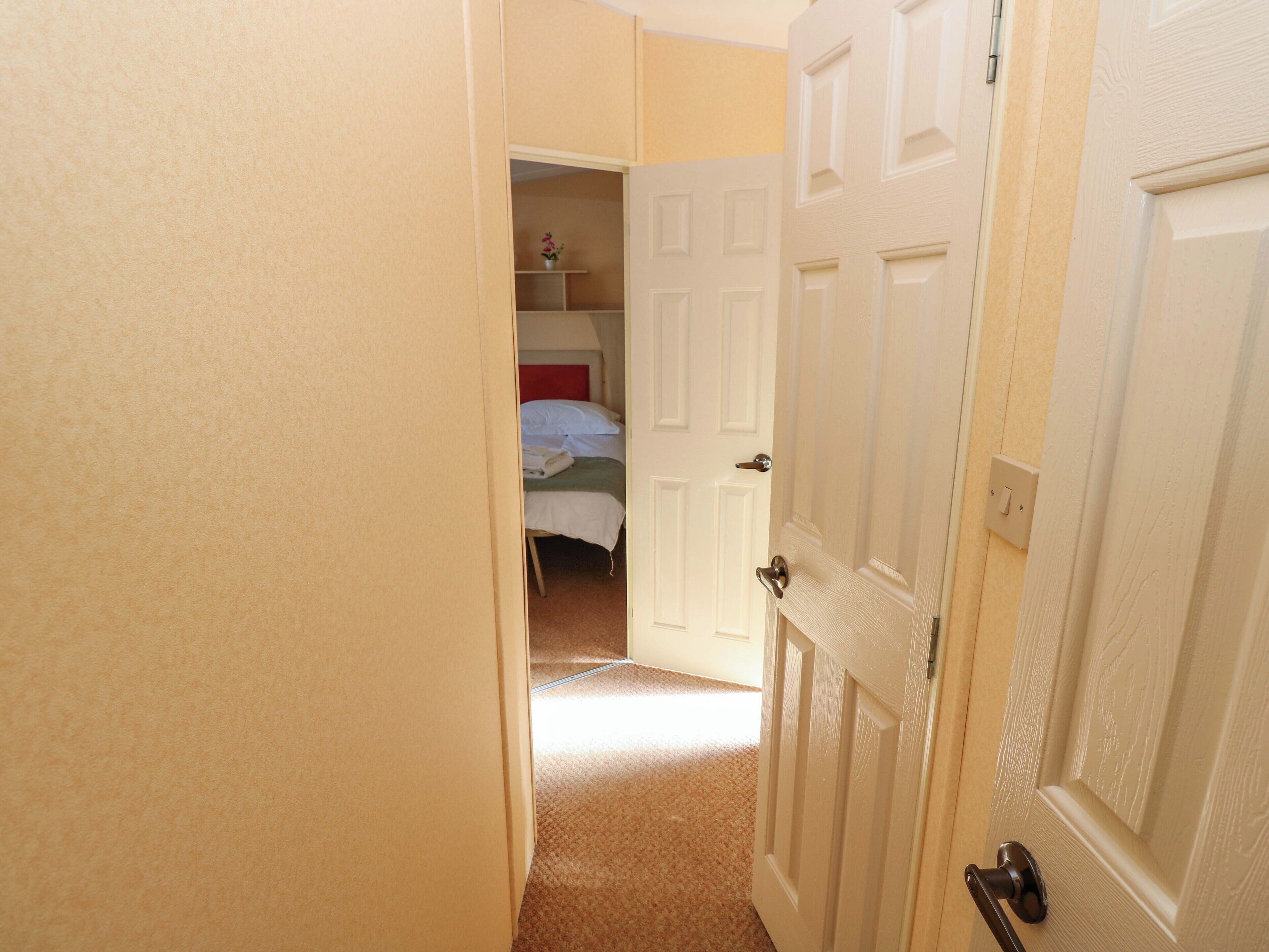 A narrow hallway leading to a bedroom with a bed, a nightstand, and a small shelf with a vase.