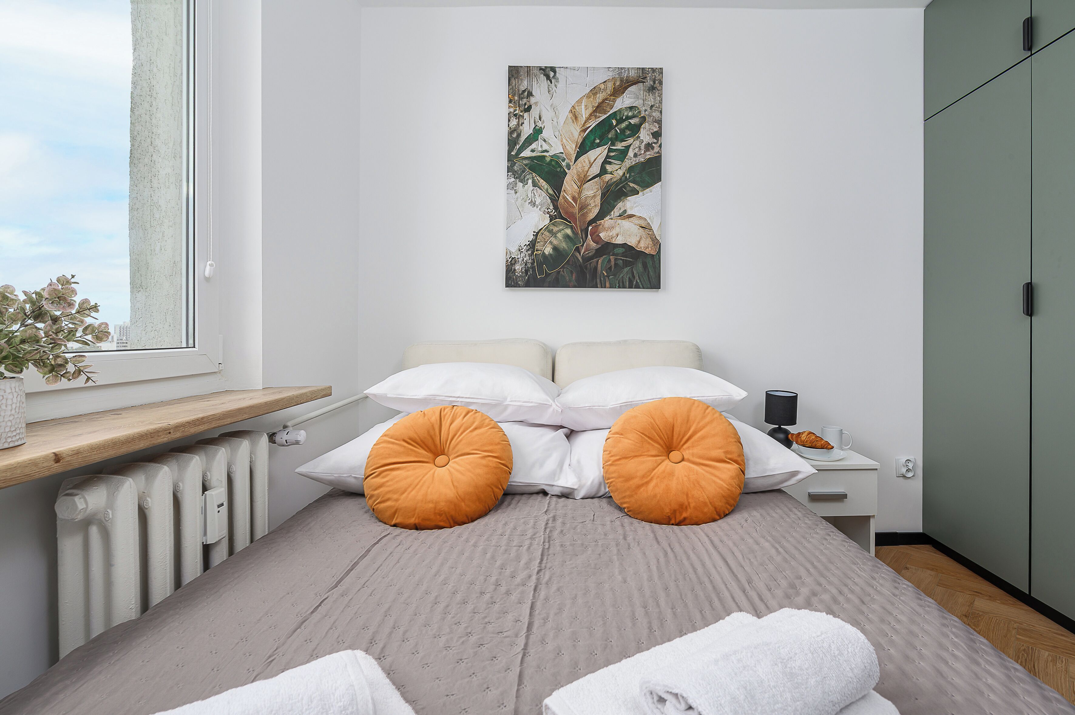 A modern bedroom with a bed, pillows, a nightstand, a radiator, a window with a plant, and a green wardrobe.