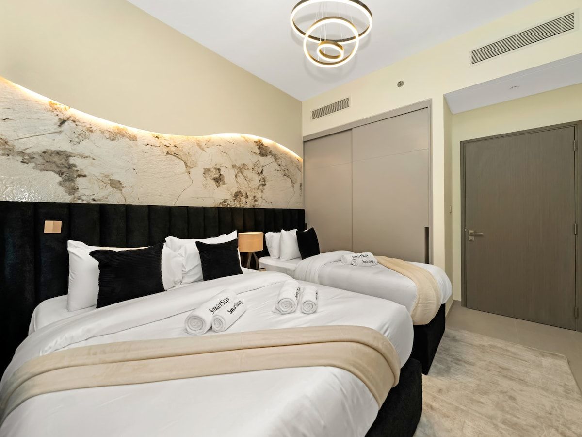 A hotel room with two beds, a marble headboard, a large mirror, a wardrobe, and a modern light fixture.