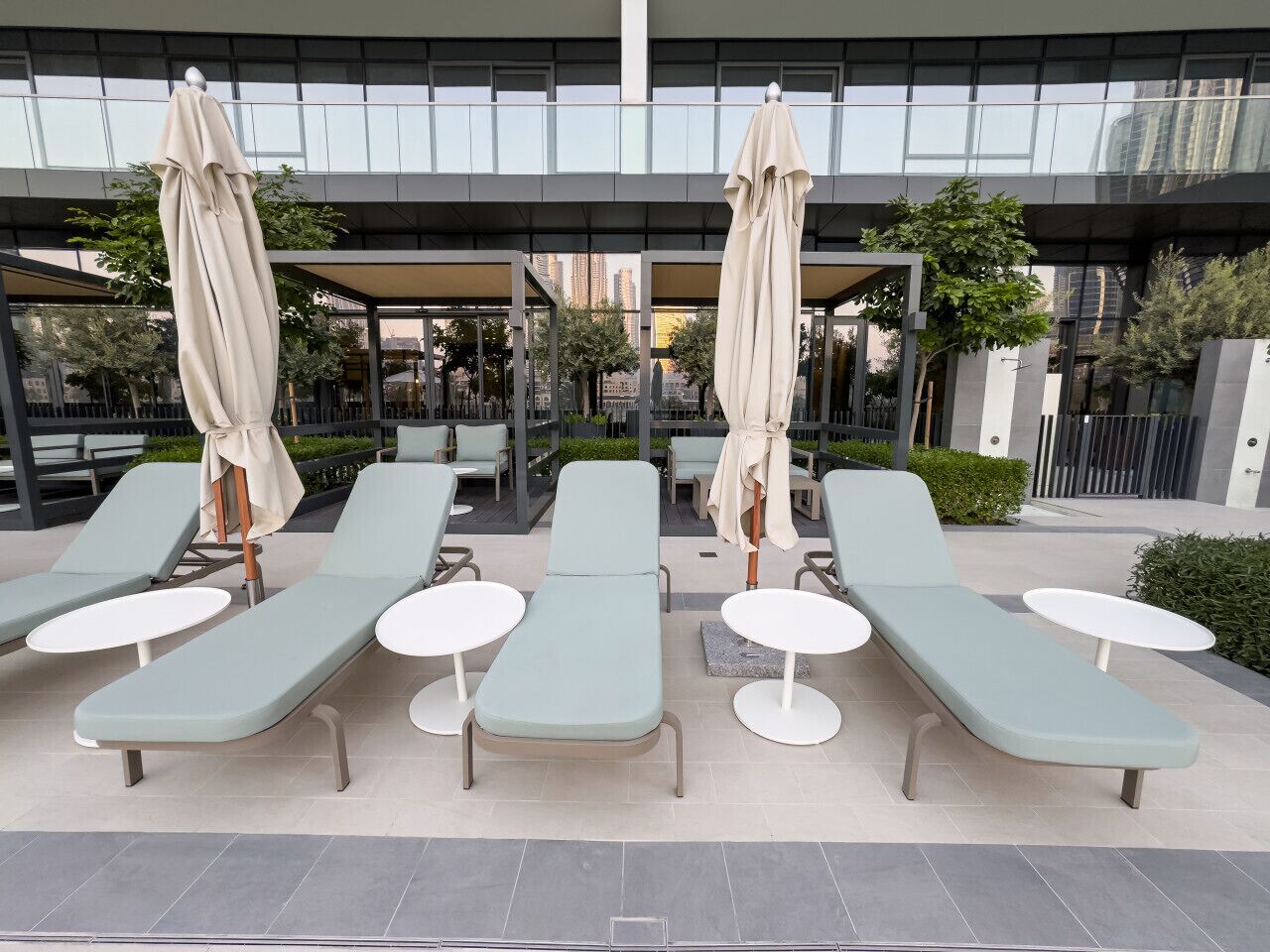 Un espace de détente extérieur moderne, avec des fauteuils bleu clair et des tables rondes blanches, chacune agrémentée d’un parasol beige.