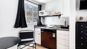 A compact kitchen with a dishwasher, microwave, and a small sink. There is a window with black curtains, a black chair, and a black table.