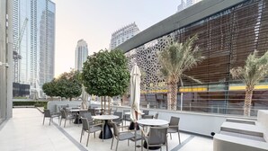 Un área de café al aire libre con mesas y sillas, rodeada de follaje y edificios modernos al fondo.