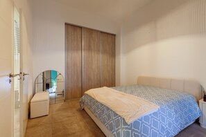 A bedroom with a bed, a small bedside table, a nightstand, a mirror, a wardrobe, and a door.