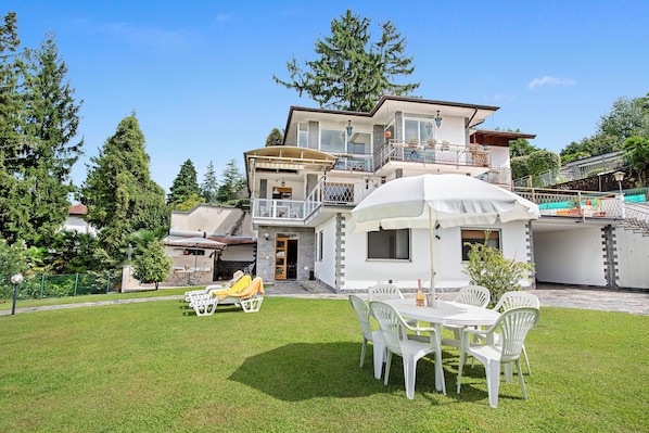 A two-story house with a balcony, a patio with a table and chairs, and a swimming pool in the background.