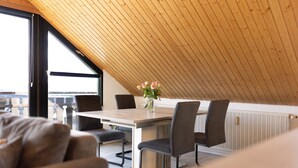 Zona pranzo con soffitto in legno, tavolo e sedie, un vaso con fiori e vista attraverso ampie finestre.