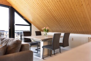 Ein Essbereich mit Holzbalkendecke, einem Tisch mit Stühlen, einer Blumenvase und Blick durch große Fenster.
