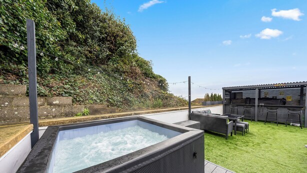 A hot tub with a stone surround, a wooden deck, and a seating area with a sofa and chairs. - ELEVATED ESCAPE - 3 bedroom house with rooftop terrace,sauna,hot tub&cinema room (Hythe)