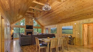 Interior acogedor de una cabaña con techo de madera, chimenea, zona de comedor y ventanas amplias que ofrecen vistas al exterior.