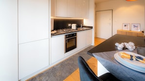 A modern kitchen with white cabinetry, a black countertop, and a built-in oven. There is a dining area with a table and chairs, and a decorative cotton plant on the table. - Emilia Suite Garden (Modena)