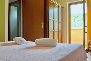 A hotel room with a bed, a folded towel, a wardrobe, and a view of greenery through a window. - Spacious three-room apartments (about 50 square meters), they are arranged as follows: <br>living room with kitchenette and double sofa bed, double bedroom, bedroom with one or two single beds and bathroom with shower.Equipped with a large covered balcon (Campiglia Marittima)
