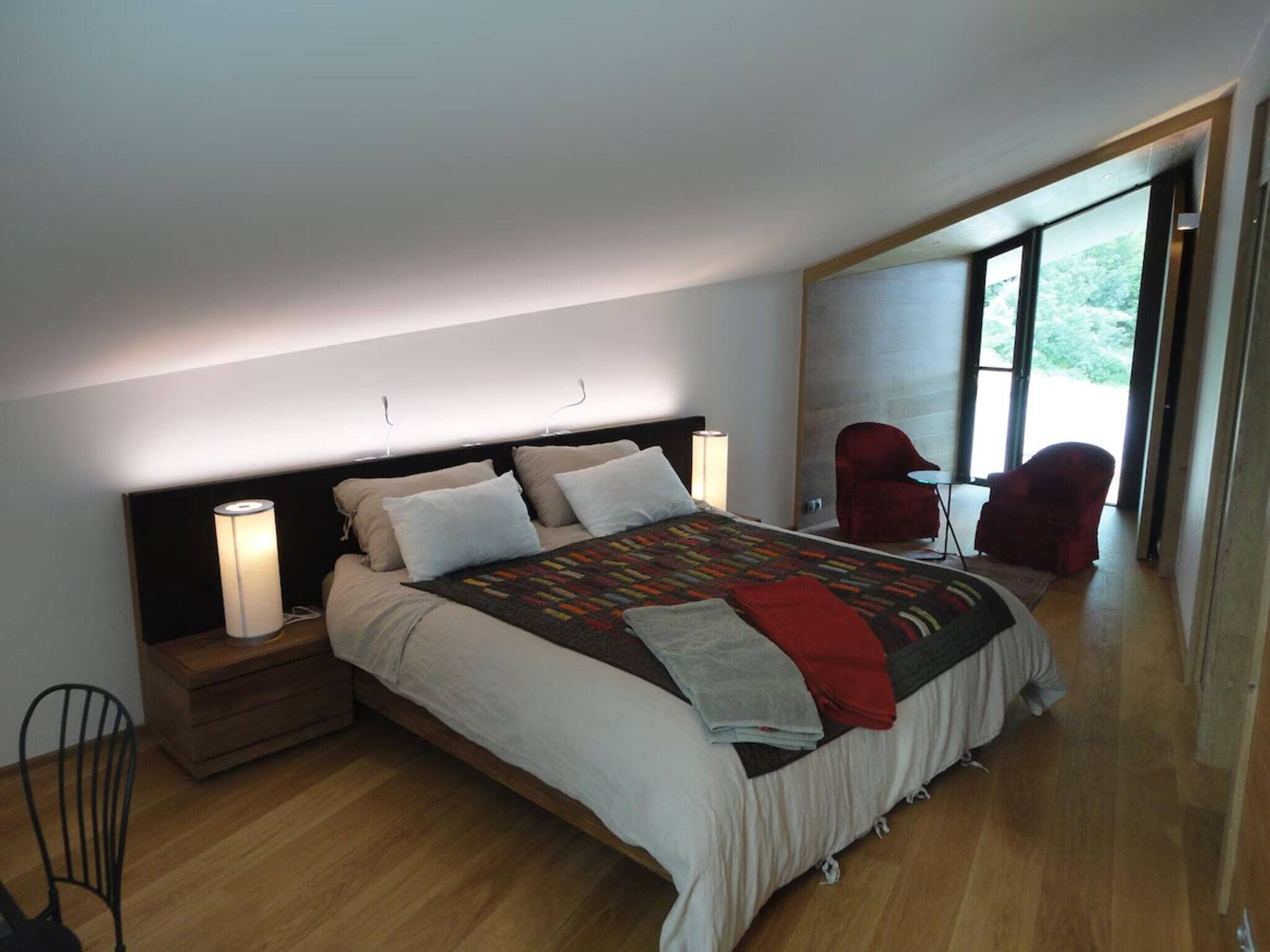 A modern bedroom with a large bed, bedside lamps, a chair, and a view of greenery outside.