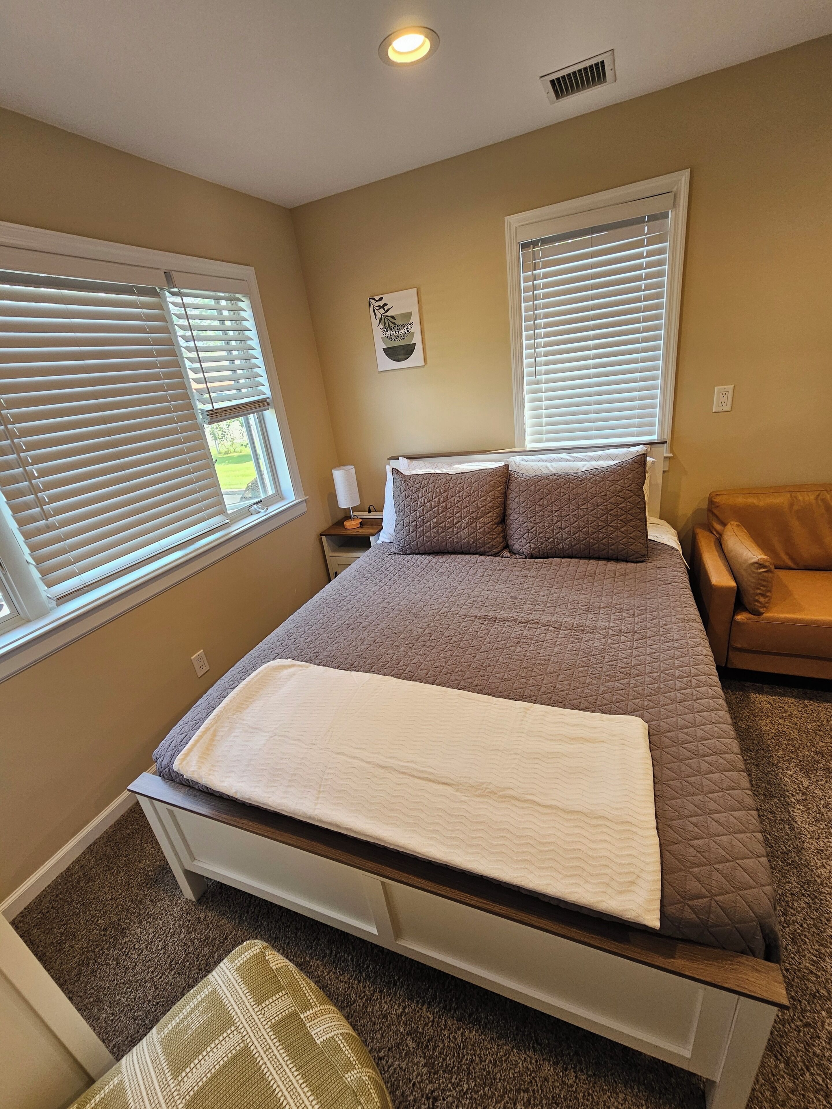 Un dormitorio con cama, dos ventanas con persianas, una mesita de noche, una lámpara, una obra de arte en la pared y un sillón de cuero marrón.
